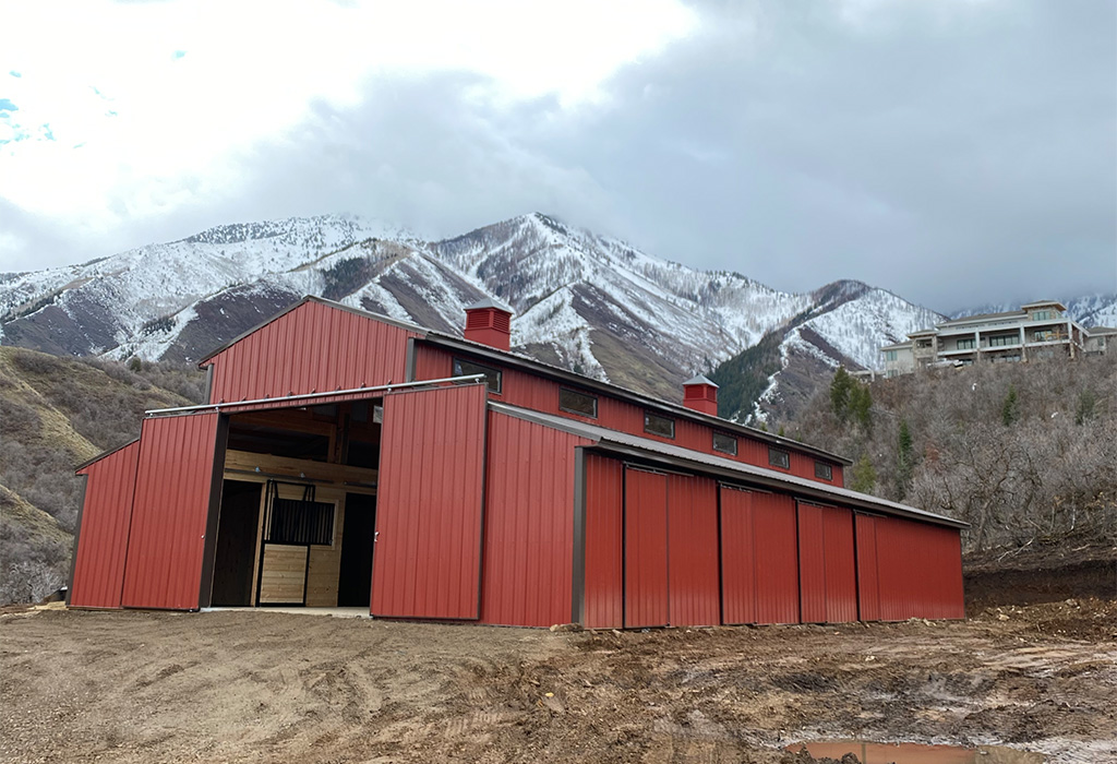 A large red horse barn with sliding doors, set against a mountainous, snow-dusted backdrop.